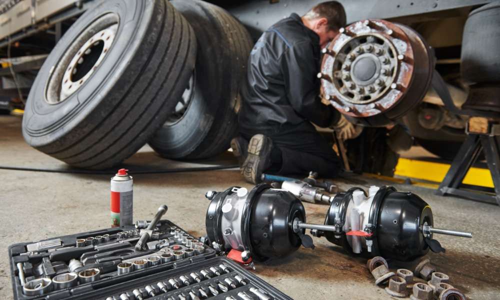 A mechanic works on a truck brake assembly in a workshop, with tools and parts spread across the floor nearby.