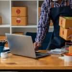 Worker holding stacked boxes while using laptop at desk, with packing supplies and shelves in small shipping workspace.