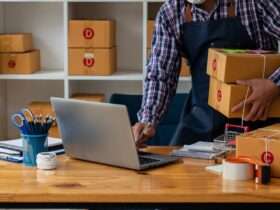 Worker holding stacked boxes while using laptop at desk, with packing supplies and shelves in small shipping workspace.