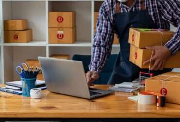 Worker holding stacked boxes while using laptop at desk, with packing supplies and shelves in small shipping workspace.
