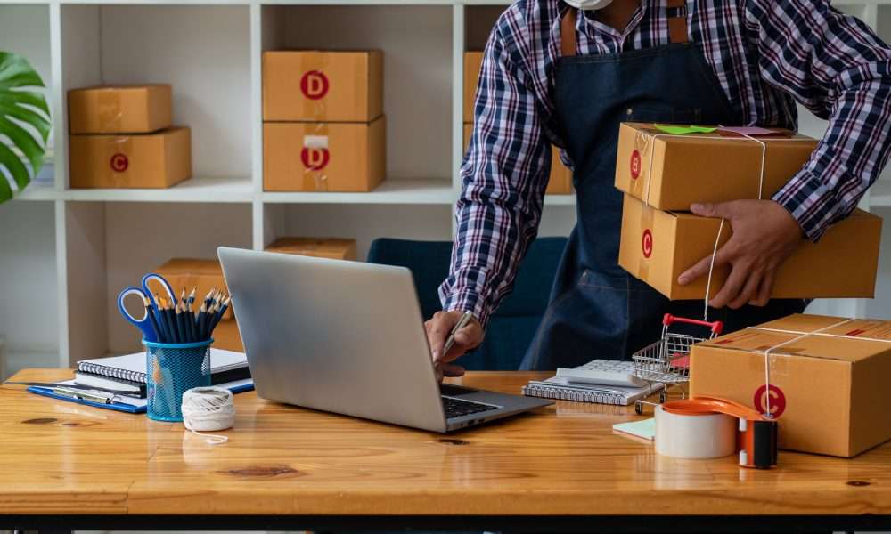 Worker holding stacked boxes while using laptop at desk, with packing supplies and shelves in small shipping workspace.