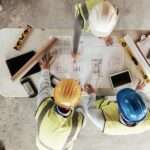 A group of construction workers reviewing blueprints on a table with various tools. They are all wearing safety gear.