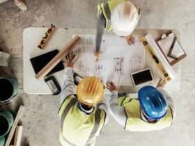 A group of construction workers reviewing blueprints on a table with various tools. They are all wearing safety gear.