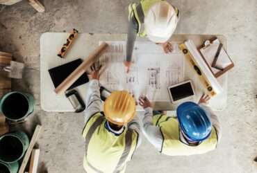 A group of construction workers reviewing blueprints on a table with various tools. They are all wearing safety gear.