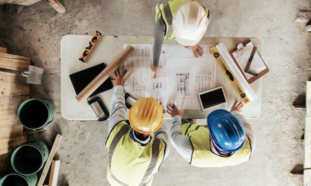 A group of construction workers reviewing blueprints on a table with various tools. They are all wearing safety gear.