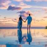 A couple stands in an infinity pool in the Caribbean and watches the sun set. The two hold each other's hands.