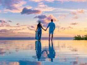 A couple stands in an infinity pool in the Caribbean and watches the sun set. The two hold each other's hands.