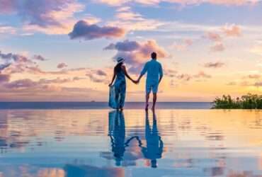 A couple stands in an infinity pool in the Caribbean and watches the sun set. The two hold each other's hands.
