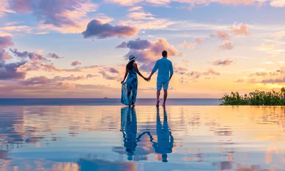 A couple stands in an infinity pool in the Caribbean and watches the sun set. The two hold each other's hands.