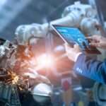 An engineer in a suit stands to the side of two robotic arms working on a piece of machinery with a tablet in his hand.