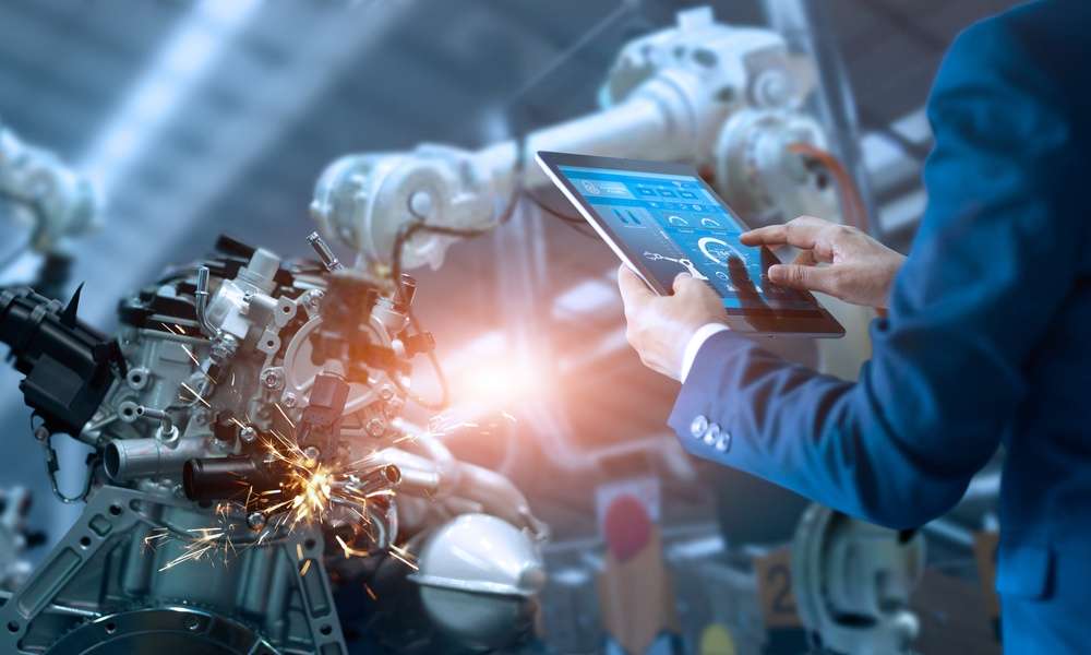 An engineer in a suit stands to the side of two robotic arms working on a piece of machinery with a tablet in his hand.