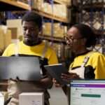 Two warehouse workers reviewing inventory using laptop and tablet, standing among shelves filled with boxes.