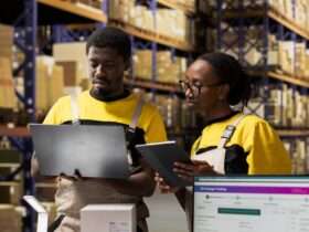 Two warehouse workers reviewing inventory using laptop and tablet, standing among shelves filled with boxes.
