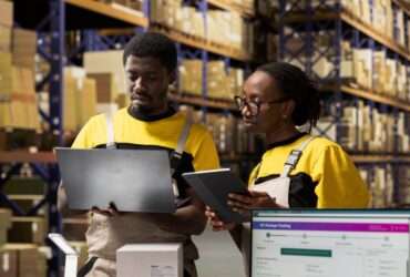 Two warehouse workers reviewing inventory using laptop and tablet, standing among shelves filled with boxes.