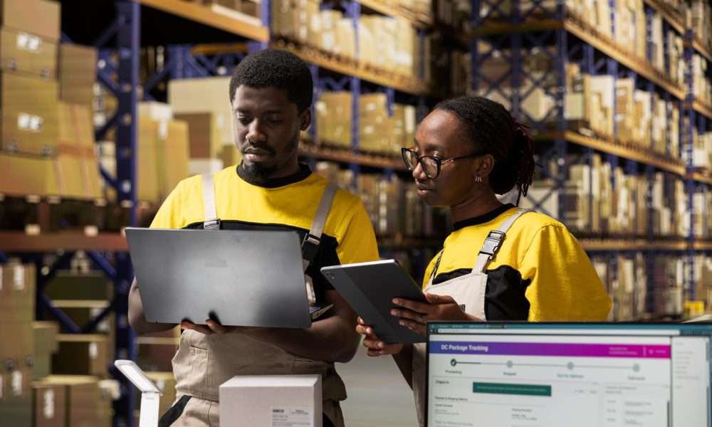 Two warehouse workers reviewing inventory using laptop and tablet, standing among shelves filled with boxes.