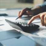 A close-up of a person using a calculator with one hand while pointing to papers on a desk with a pencil in the other hand.