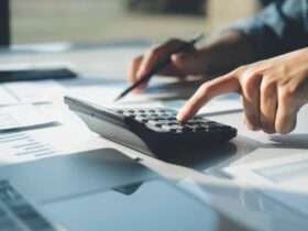 A close-up of a person using a calculator with one hand while pointing to papers on a desk with a pencil in the other hand.