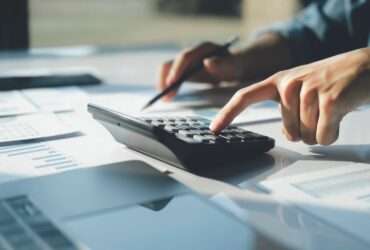 A close-up of a person using a calculator with one hand while pointing to papers on a desk with a pencil in the other hand.