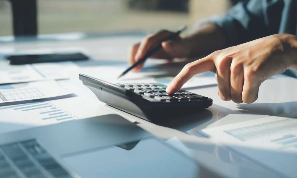 A close-up of a person using a calculator with one hand while pointing to papers on a desk with a pencil in the other hand.