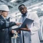 A worker in a hard hat and safety glasses points to a laptop a technician holds while standing in a factory.