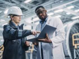 A worker in a hard hat and safety glasses points to a laptop a technician holds while standing in a factory.