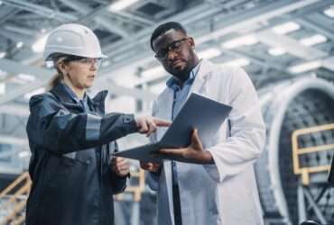 A worker in a hard hat and safety glasses points to a laptop a technician holds while standing in a factory.