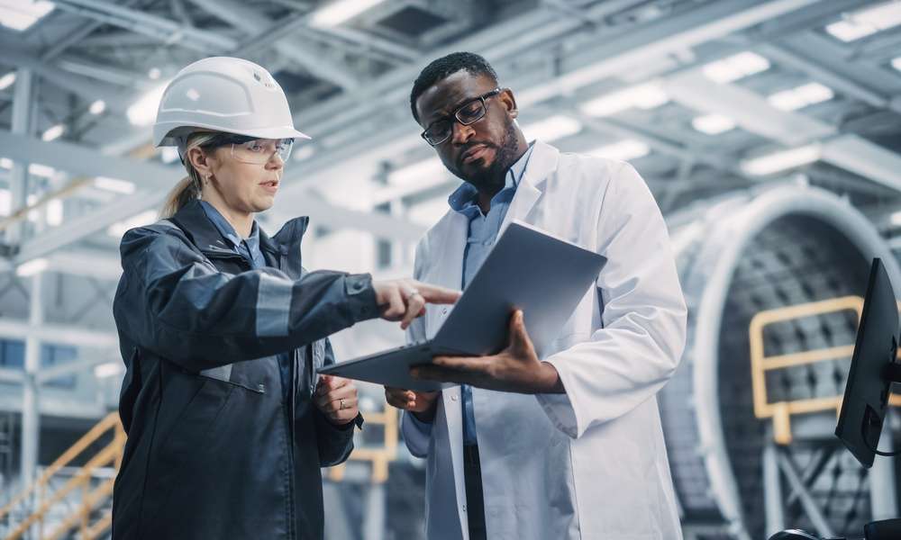 A worker in a hard hat and safety glasses points to a laptop a technician holds while standing in a factory.