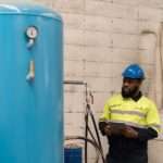 A worker in safety gear inspects a large vertical blue air tank with a pressure gauge inside an industrial facility.