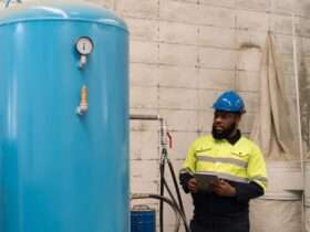 A worker in safety gear inspects a large vertical blue air tank with a pressure gauge inside an industrial facility.