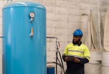 A worker in safety gear inspects a large vertical blue air tank with a pressure gauge inside an industrial facility.