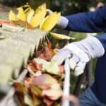 A person wearing a blue sweatshirt and gloves clears leaves and debris from a clogged roof gutter outdoors.