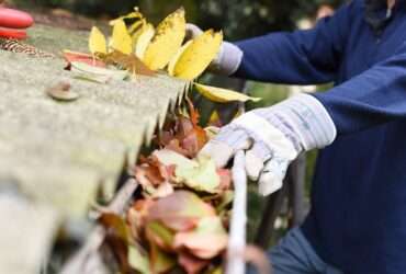 A person wearing a blue sweatshirt and gloves clears leaves and debris from a clogged roof gutter outdoors.
