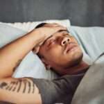 A young man lies in bed with the sheets pulled up to his chest. He has his eyes closed with his hand over his forehead.