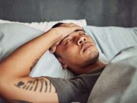 A young man lies in bed with the sheets pulled up to his chest. He has his eyes closed with his hand over his forehead.