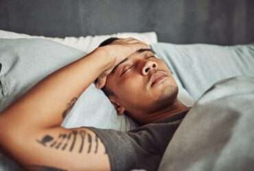 A young man lies in bed with the sheets pulled up to his chest. He has his eyes closed with his hand over his forehead.