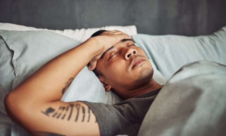 A young man lies in bed with the sheets pulled up to his chest. He has his eyes closed with his hand over his forehead.