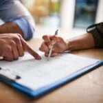 A close-up of a hand pointing at a line on a piece of paper on a wooden desk, while another hand holds a pen.