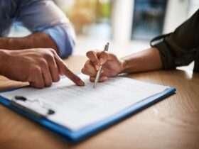 A close-up of a hand pointing at a line on a piece of paper on a wooden desk, while another hand holds a pen.