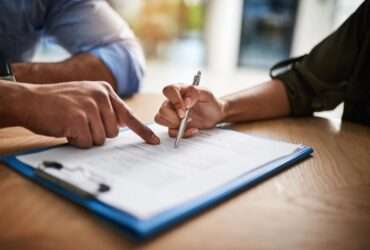 A close-up of a hand pointing at a line on a piece of paper on a wooden desk, while another hand holds a pen.