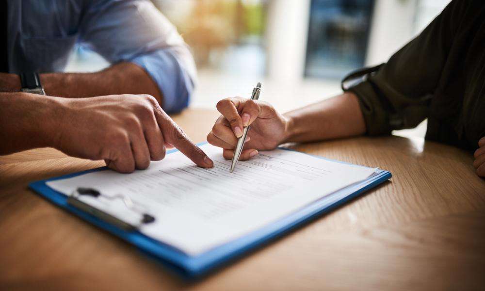 A close-up of a hand pointing at a line on a piece of paper on a wooden desk, while another hand holds a pen.
