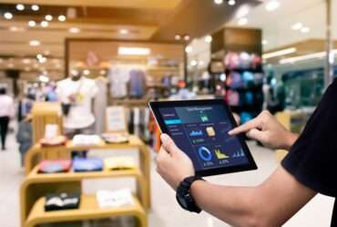 A manager holding a tablet with data and figures about store management. Behind the tablet is a store with apparel.