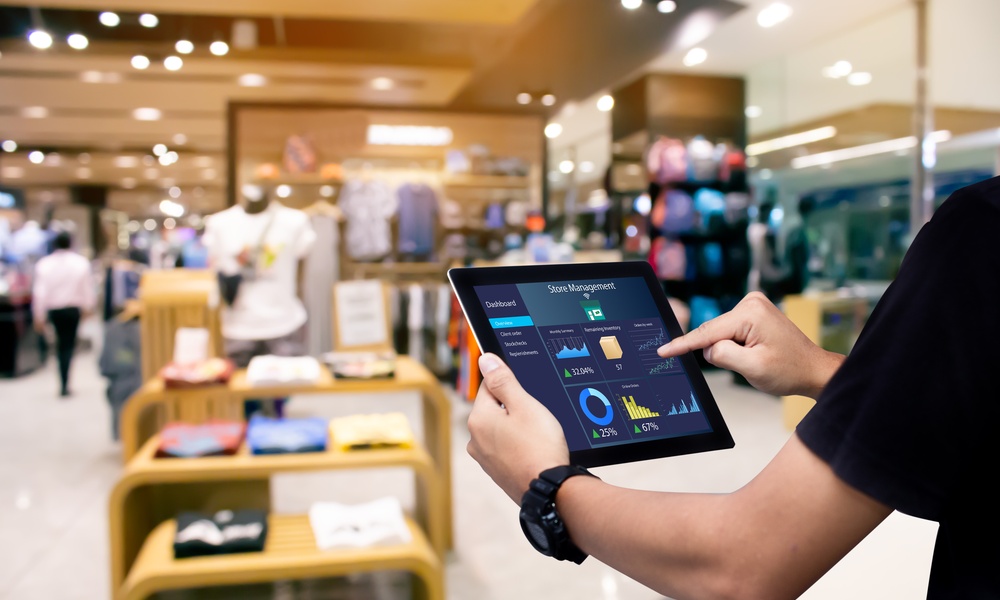 A manager holding a tablet with data and figures about store management. Behind the tablet is a store with apparel.