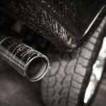 A black truck’s exhaust pipe, positioned behind a rugged tire, with dirt splattering the pipe and the body of the truck.