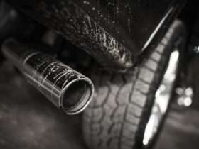 A black truck’s exhaust pipe, positioned behind a rugged tire, with dirt splattering the pipe and the body of the truck.