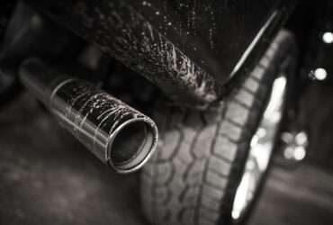 A black truck’s exhaust pipe, positioned behind a rugged tire, with dirt splattering the pipe and the body of the truck.