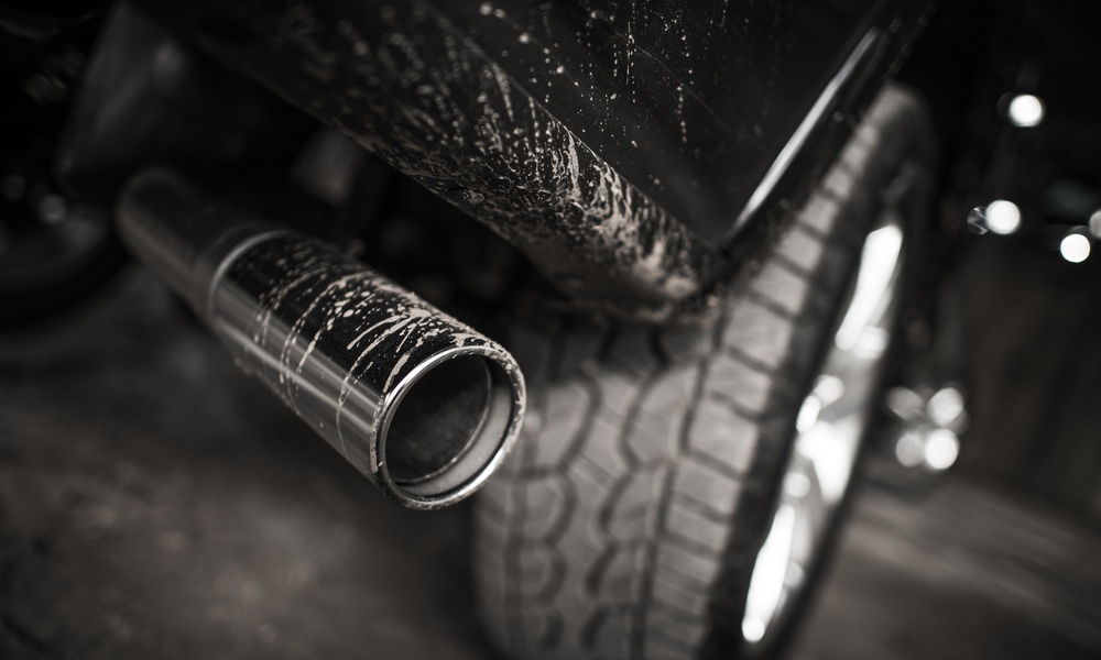 A black truck’s exhaust pipe, positioned behind a rugged tire, with dirt splattering the pipe and the body of the truck.