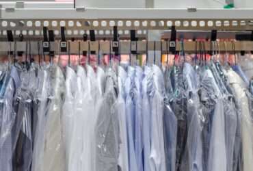 Several dress shirts are hanging on hangars and are placed on a rack. Each shirt has a plastic cover on them.