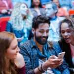 Three attendees at a crowded conference smile while looking at a smartphone as others sit and talk in the background.