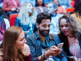 Three attendees at a crowded conference smile while looking at a smartphone as others sit and talk in the background.