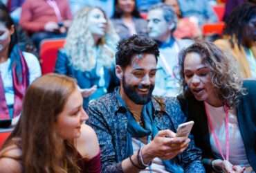 Three attendees at a crowded conference smile while looking at a smartphone as others sit and talk in the background.